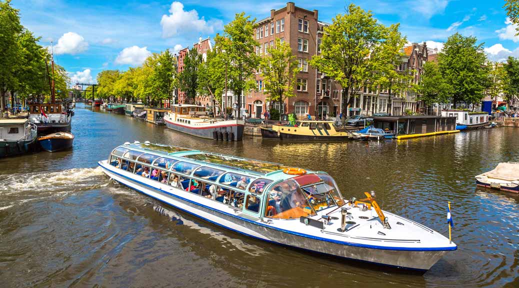 Canals of Amsterdam with tourist boat with people in the background green tress and houses.